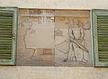 Relief mit griechischem Motiv an Hausfassade zwischen Fensterläden